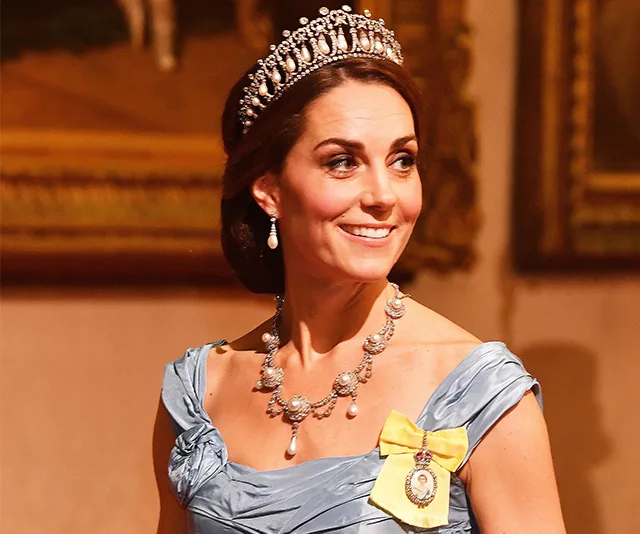 A woman in a blue gown and a jeweled tiara, smiling, with portraits in the background.