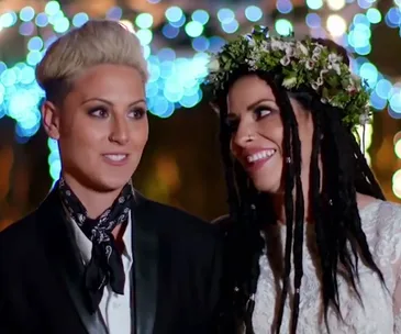 A couple smiles at each other at their wedding, one in a suit with a scarf, the other in a dress with flower crown.