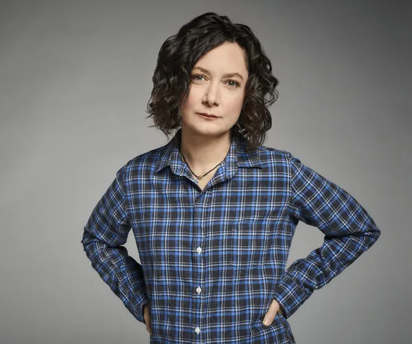 Sara Gilbert posing in a blue plaid shirt with hands on hips against a gray background.