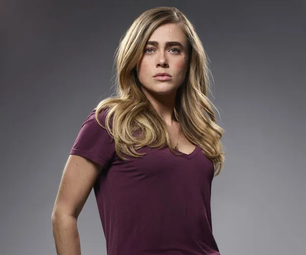 Actress in a maroon top with long blonde hair, against a gray background.