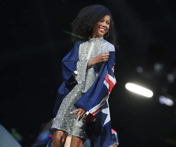 A performer wrapped in an Australian flag smiles on stage, wearing a sparkly silver dress.