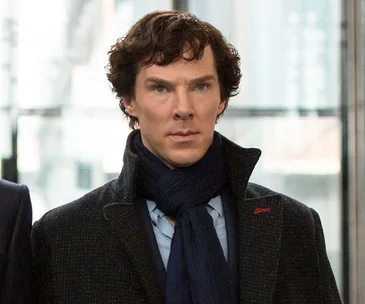 Sherlock Holmes, with curly hair, wearing a dark coat and scarf, looks on intently.