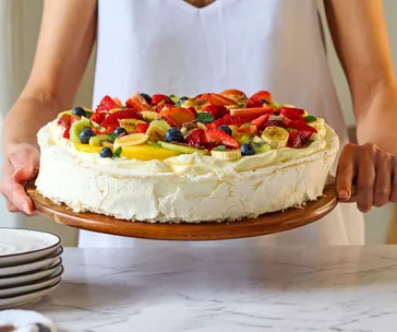 A person holding a pavlova topped with sliced fruits like strawberries, kiwi, and blueberries on a wooden platter.