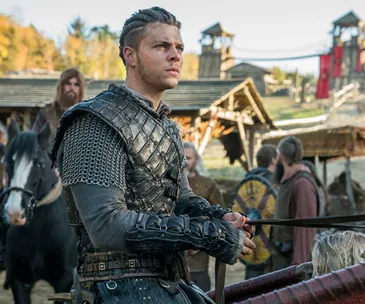 Young man in Viking armor stands with a determined expression in a medieval village setting.