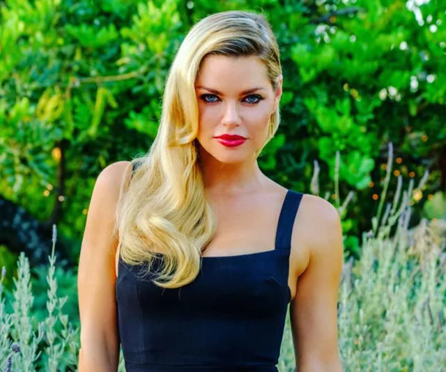 Blonde woman in black dress with lush green background.