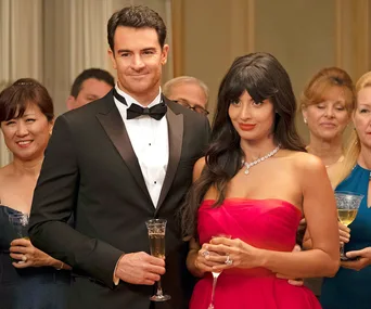 A couple in formal attire holding champagne flutes at a social event, surrounded by other elegantly dressed guests.