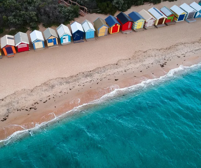 Aerial view of colorful beach huts along a sandy shore with turquoise ocean waves in Melbourne.
