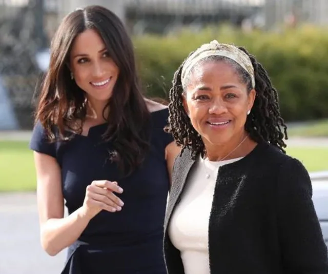 Women smiling outdoors, one in a dark dress and the other in a sweater, with greenery in the background.