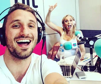 Two radio hosts smiling and wearing headphones in a studio. One is taking a selfie; the other gestures energetically.