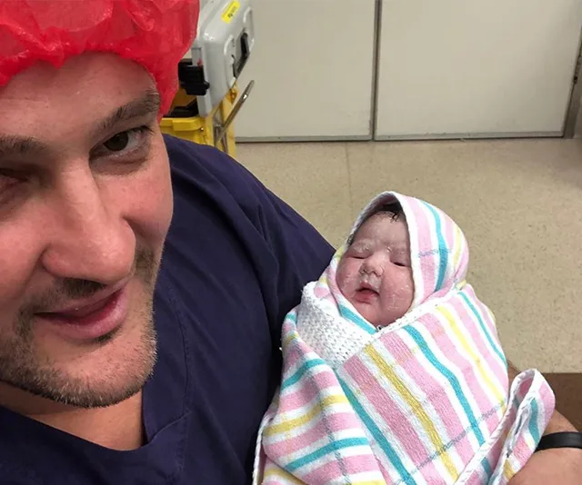 A person wearing a red hair net holds a newborn wrapped in a striped blanket in a hospital setting.