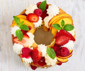 Fruit-adorned holiday bundt cake with strawberries, peaches, raspberries, mint, and whipped cream on a light wooden table.