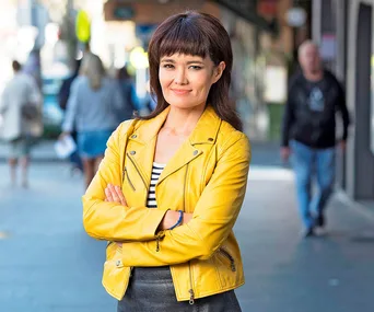 A person in a yellow jacket smiles confidently with arms crossed on a bustling street.
