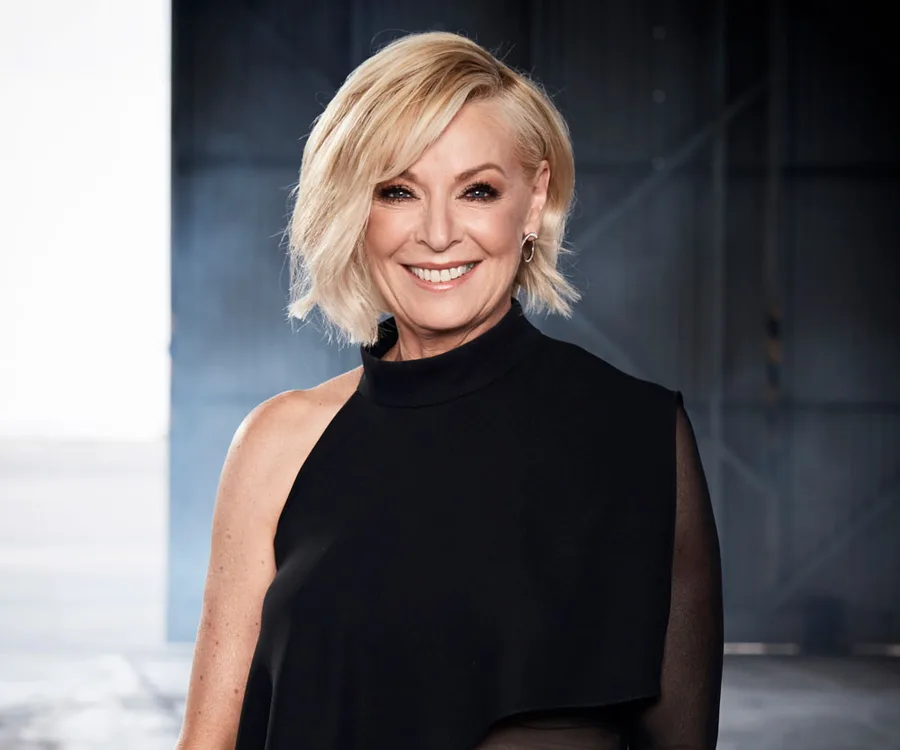 Liz Hayes smiling in a black one-shoulder outfit with a blurred industrial background.