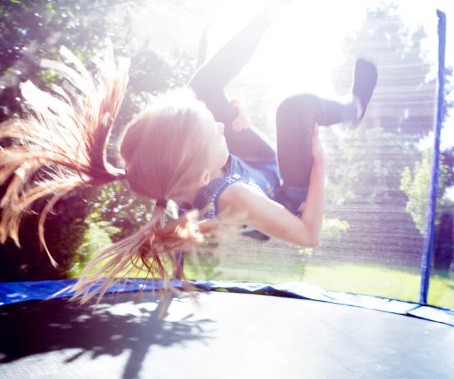 Girl with long hair jumps on a trampoline outdoors on a sunny day, captured mid-flip with a blurred motion effect.