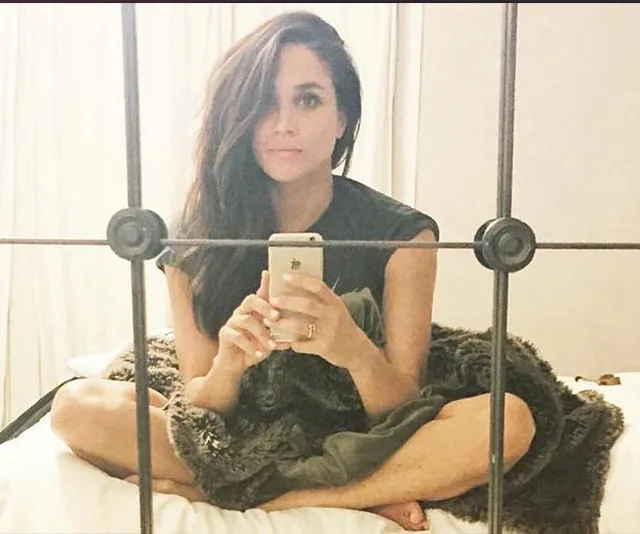 A woman sitting cross-legged on a bed takes a selfie in a mirror, wrapped in a blanket.