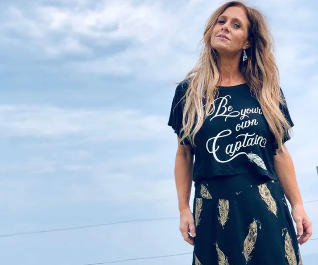 Kasey Chambers in a black shirt with "Be your own captain" text, standing outside with a cloudy sky background.