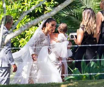 Bride in a lace gown and veil holds a child at an outdoor wedding with guests nearby.