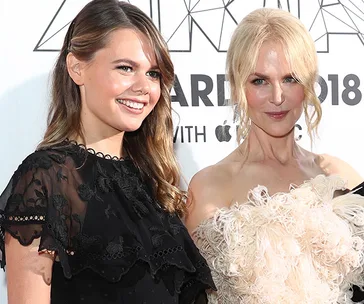 Two women smiling in glamorous outfits on a red carpet event. One wears a black dress, the other a light-colored feathered gown.