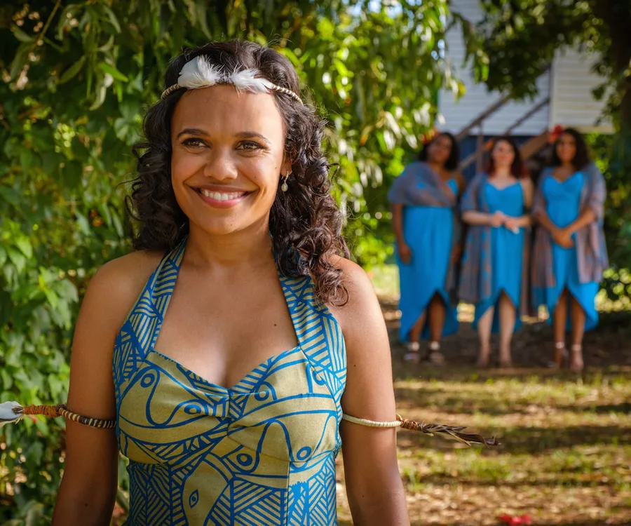 A woman in a patterned dress with a feather headband smiles outdoors, with three women in blue dresses in the background.