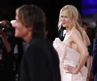 A woman in a pale dress poses confidently on the red carpet, with a man blurred in the foreground.