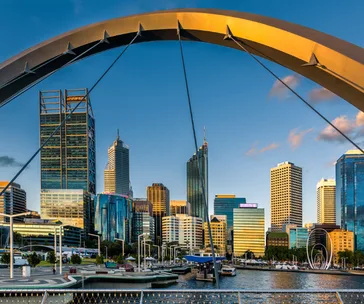 Perth skyline at sunset with modern skyscrapers and a prominent bridge arch framing the city, reflecting on the water below.