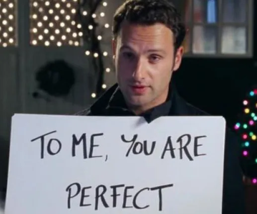 Man holding a sign reading "To me, you are perfect" in a scene with festive lights in the background.