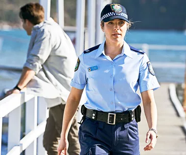 Policewoman in uniform walking confidently on a dock, with a man leaning on the railing in the background.