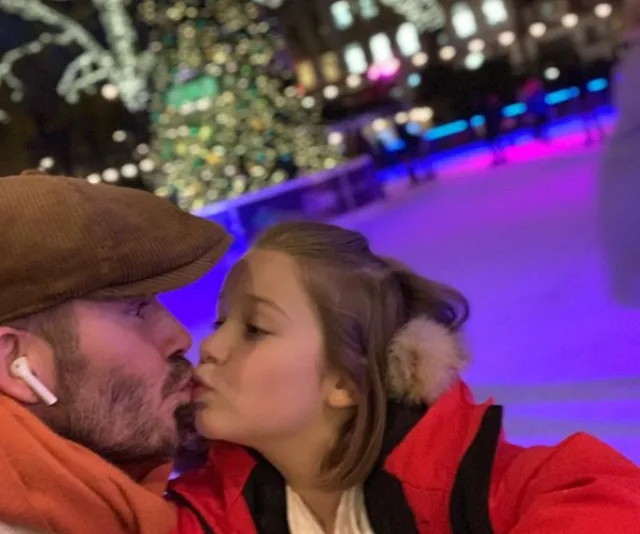 Man and child in winter clothes share a kiss at a festive outdoor ice rink with a lit Christmas tree in the background.