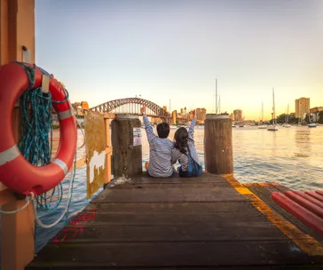 Two children in matching striped shirts sit on a dock with arms raised, overlooking Sydney Harbour Bridge at sunset.