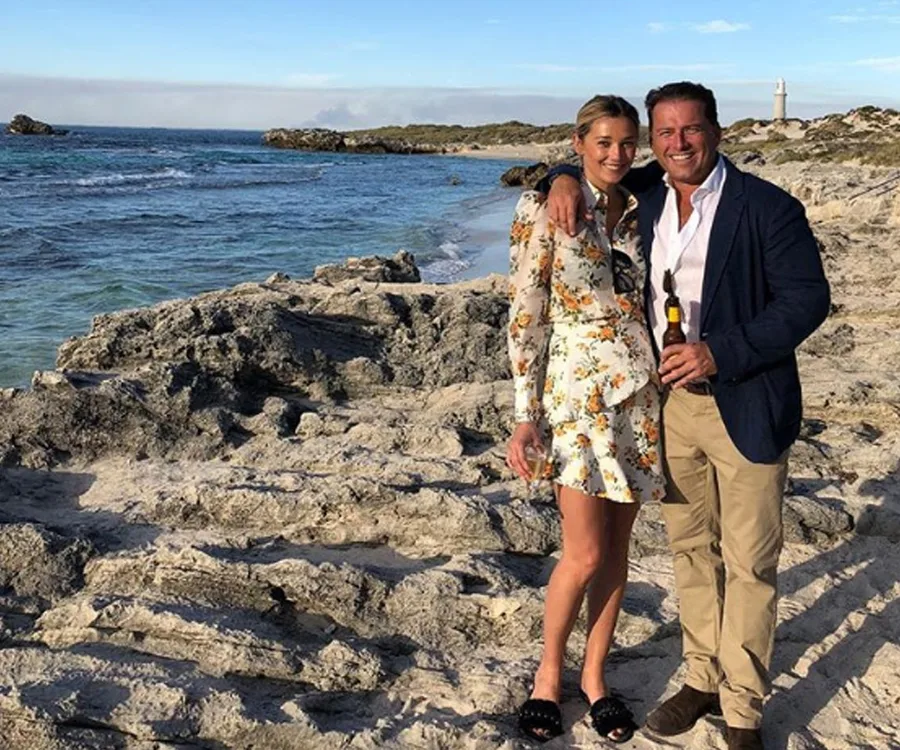 A couple posing on a rocky beach with a lighthouse in the background, both smiling and holding drinks.