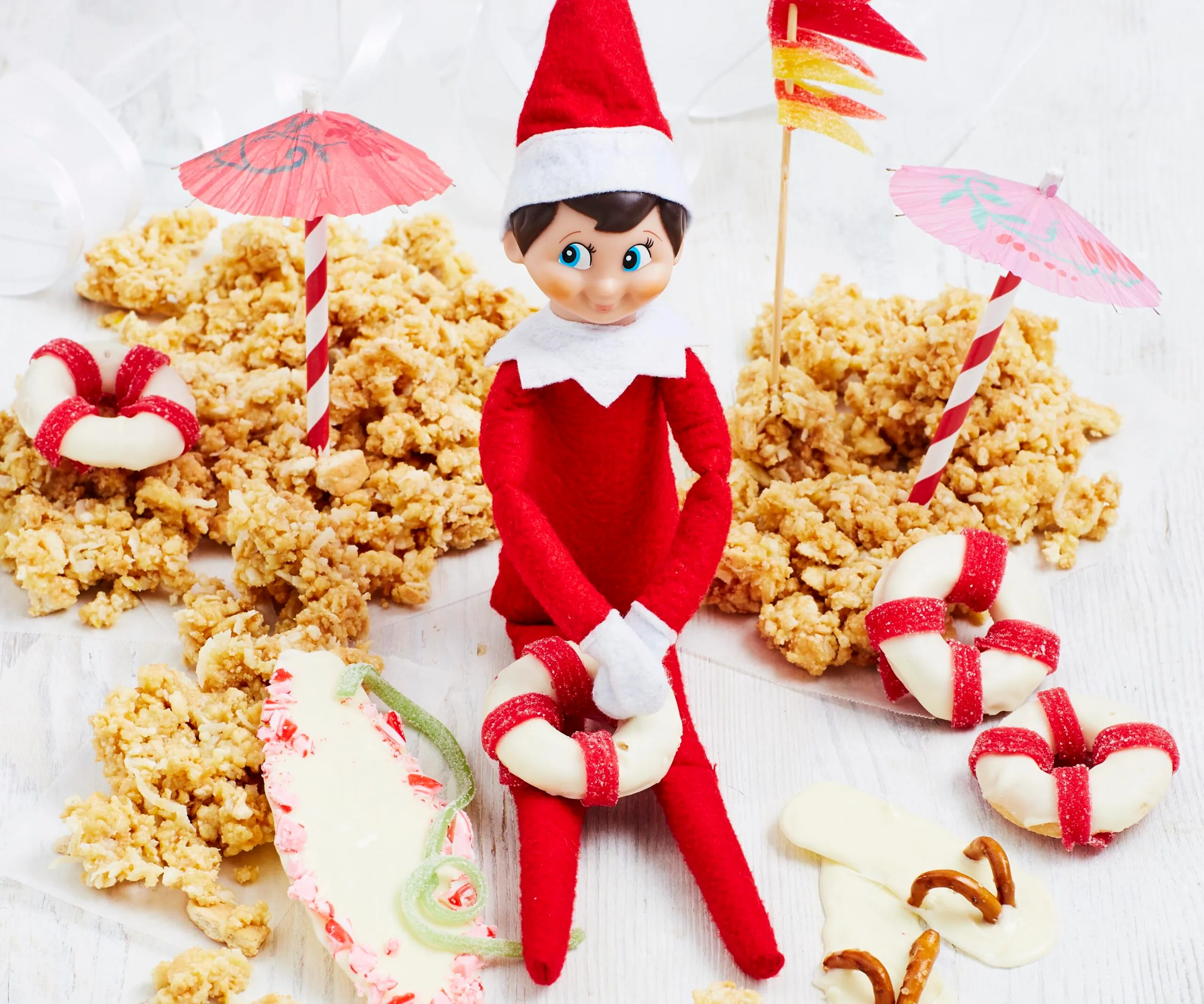 Elf on the Shelf sitting on a crunchy beach scene with umbrella, candy rings, and pretzel flip-flops.