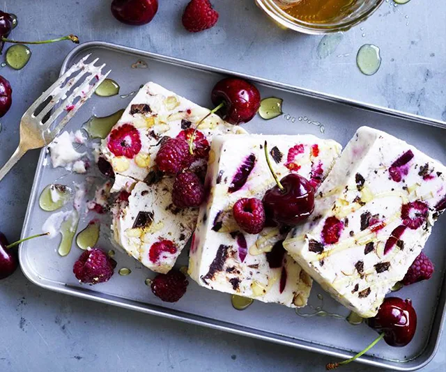 Slices of frozen nougat with raspberries, cherries, and nuts on a tray, garnished with cherries and honey drizzle.