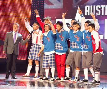Host interviewing a dance group in blue and red outfits on the "Australia's Got Talent" stage.