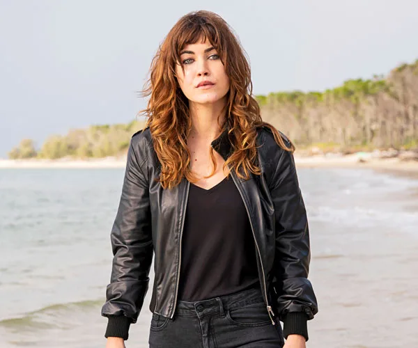 A woman with long hair in a black jacket stands on a beach with trees in the background.