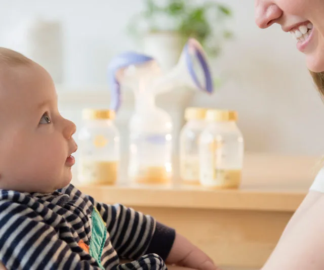 Baby looking at a smiling person with a breast pump and milk bottles on a table in the background.