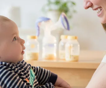 Baby looking at a smiling person with a breast pump and milk bottles on a table in the background.