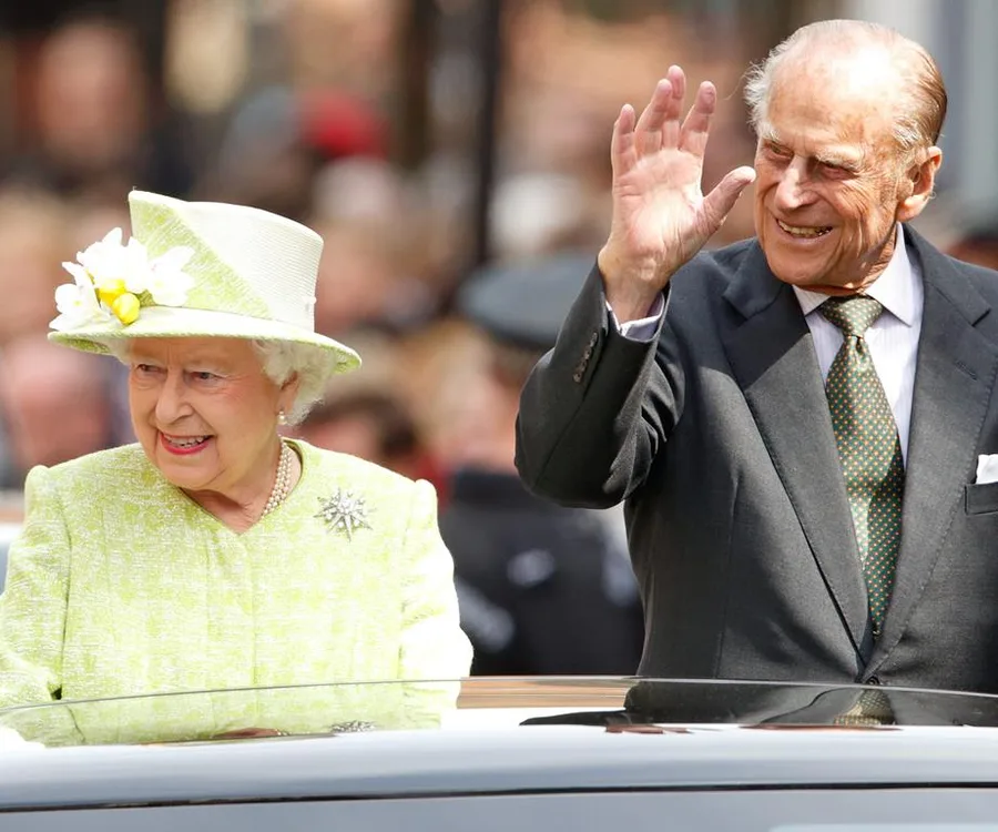 Two elderly people in formal attire, one waving, standing in an open-top vehicle during a public event.