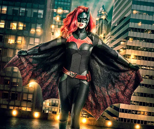 Batwoman in black and red suit with cape open, standing on rooftop with city buildings in the background.