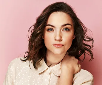 Woman with wavy brown hair in a light sequin blouse against a pink background.
