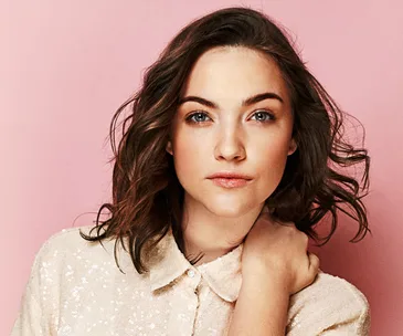 Woman with wavy brown hair in a light sequin blouse against a pink background.