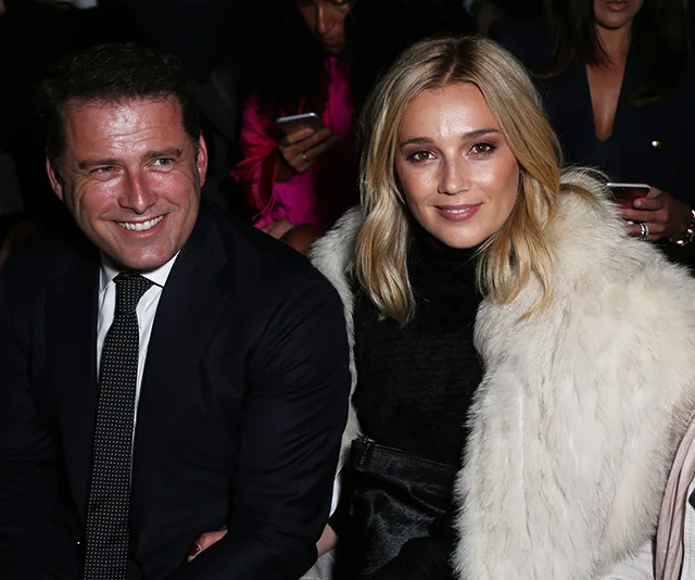 A smiling man in a suit and a woman in a fur coat sit together at an event, with people in the background using phones.