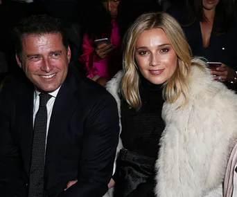 A smiling man in a suit and a woman in a fur coat sit together at an event, with people in the background using phones.