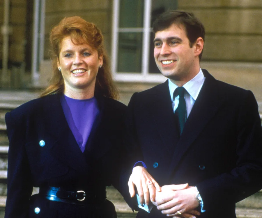 A couple smiling and holding hands, dressed in formal attire, standing outside a building.