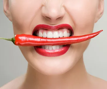 Woman with bright red lips holding a red chili pepper between her teeth, smiling.