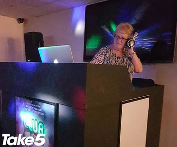 A person DJing in a dimly lit room, wearing headphones, with colorful lights and Take5 logo visible.