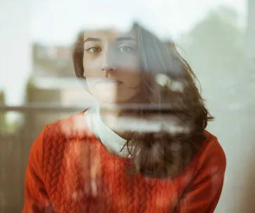 Woman with long hair in red sweater looking through a window, reflecting a contemplative mood.