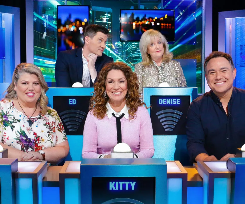 Five people sit at a quiz show panel desk with name displays reading Ed, Denise, and Kitty. The set is brightly lit.