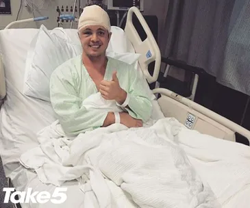 Patient in hospital bed with head bandage smiling and giving thumbs up. Wearing hospital gown, branded "Take 5" visible.