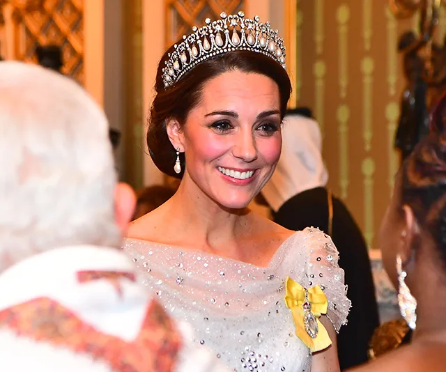 Duchess wearing a shimmering gown and diamond tiara at a formal event, smiling warmly in conversation.