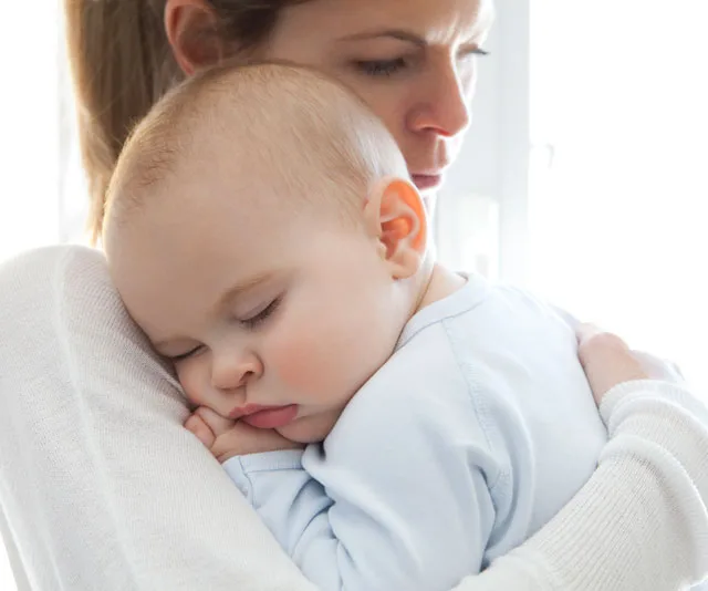 A mother gently holds her peacefully sleeping baby on her shoulder in a bright room.